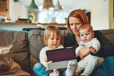 两个婴儿和一个女人坐在沙发上，一边抱着婴儿，一边看着平板电脑